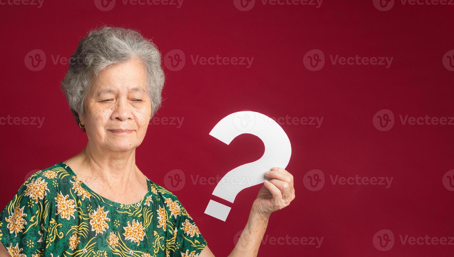 An elderly Asian woman with short gray hair holding a white question mark paper with a smile while standing on a red background photo