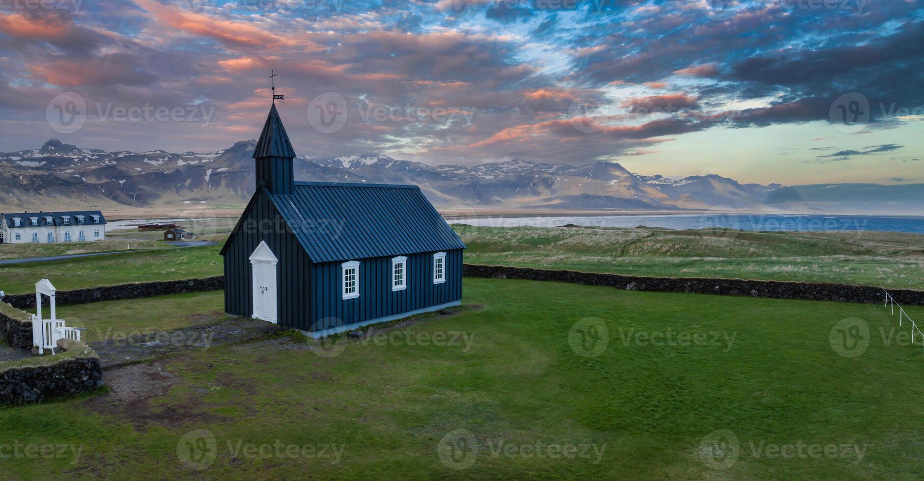 A stunning aerial view of a black church in Iceland, surrounded by a stone wall and grassy field, with mountains and a body of water in the background. photo
