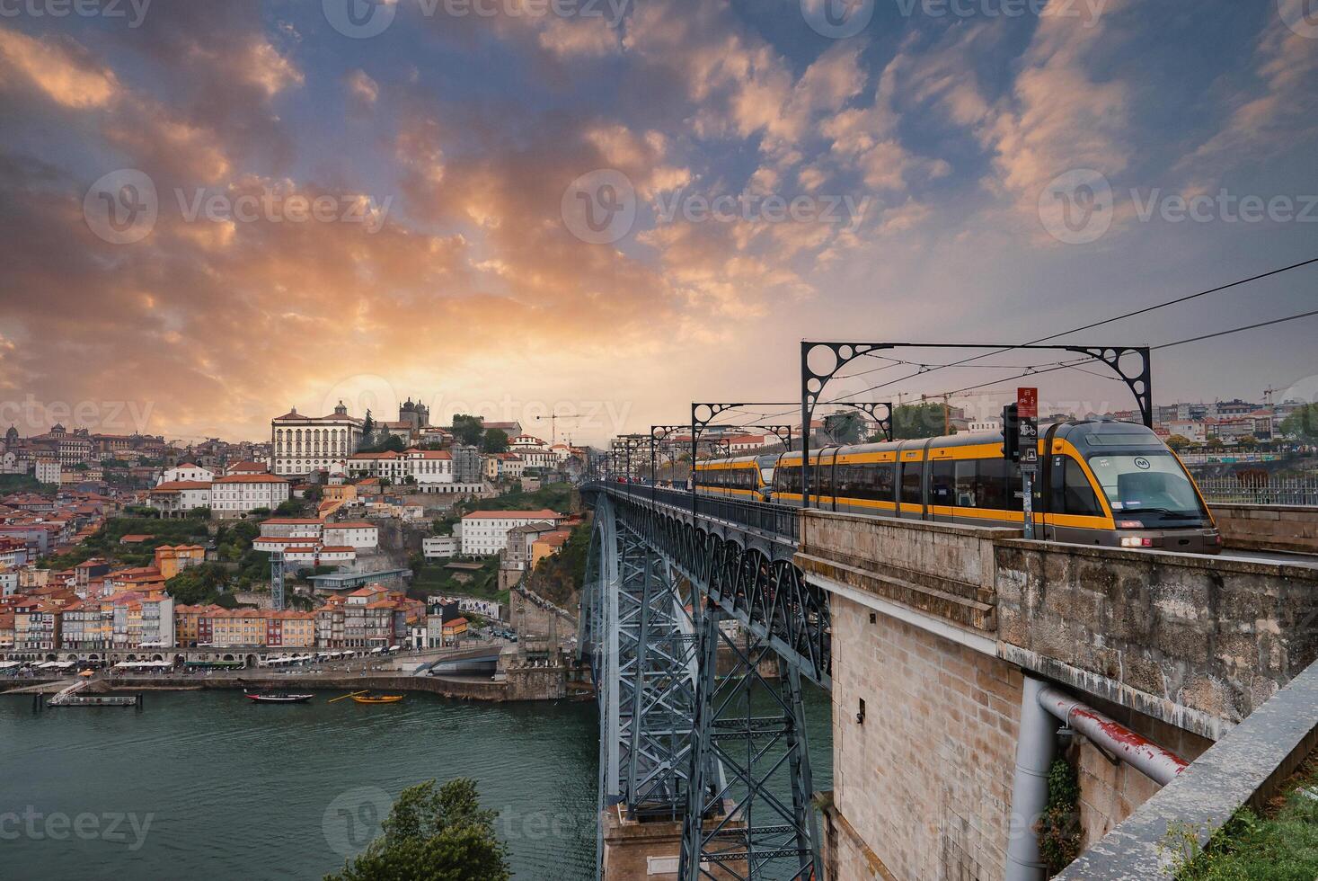 A modern tram crosses the iconic Dom Luis I Bridge over the Douro River in Porto, Portugal. The sunset illuminates historic buildings and rooftops, blending charm and infrastructure. photo