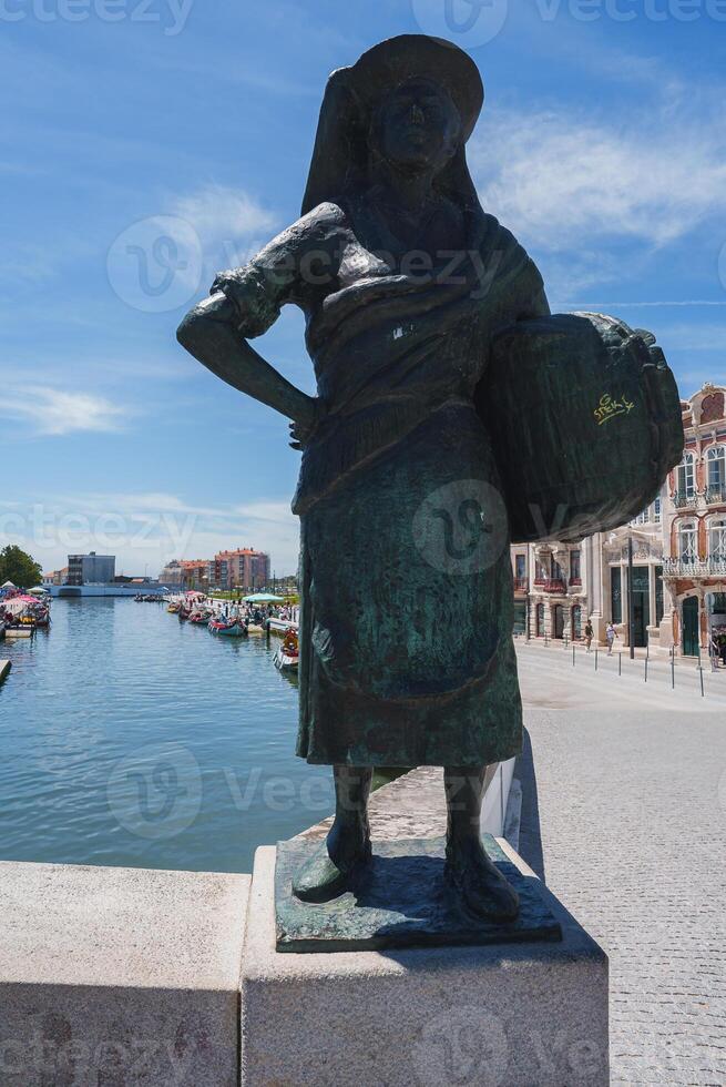 Bronze statue of a woman in traditional attire holding a basket by a canal in Aveiro, Portugal. The scene features colorful buildings, boats, and a clear blue sky. photo