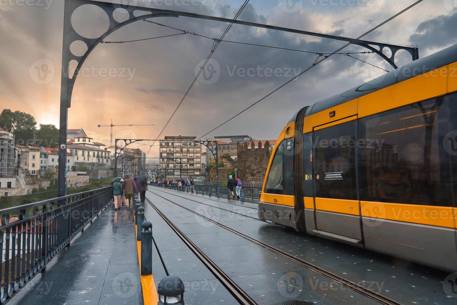 A tram traverses the iconic Dom Luis I Bridge in Porto, Portugal, during a dramatic sunset. The wet surface and historic buildings create a reflective, atmospheric scene. photo