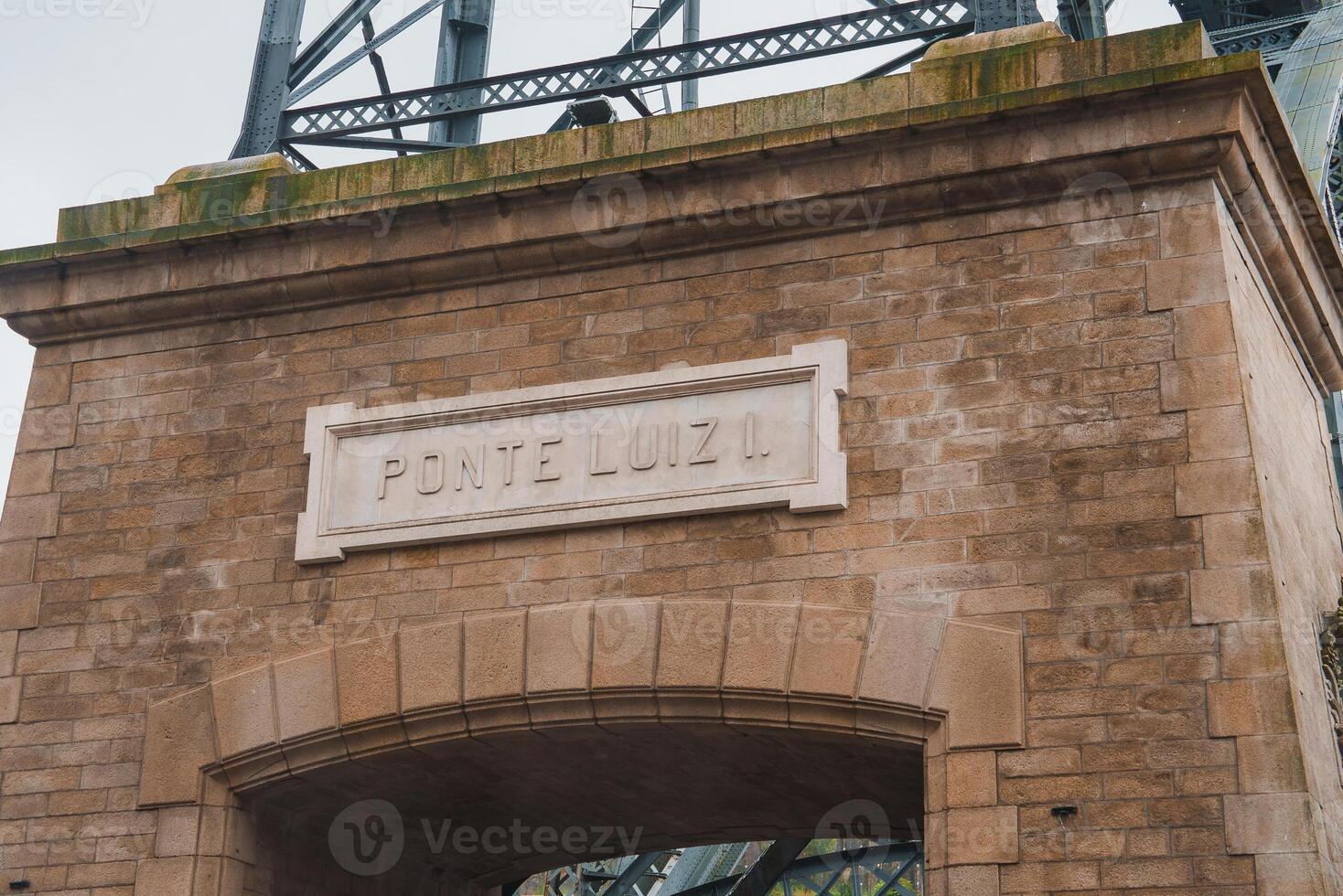 Detailed view of the stone archway and steel elements of Ponte Luiz I in Porto, Portugal. The light brown stone bricks and white plaque highlight its architectural significance. photo