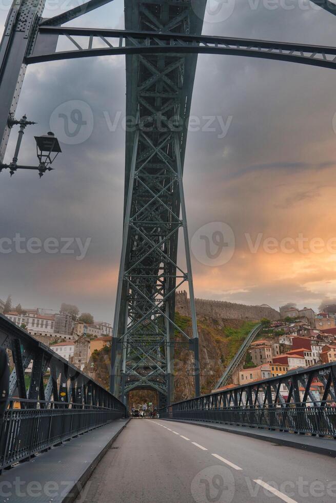View from the lower deck of the Dom Luis I Bridge in Porto, Portugal, showcasing intricate ironwork, a dramatic sunset sky, and vibrant hillside architecture. photo