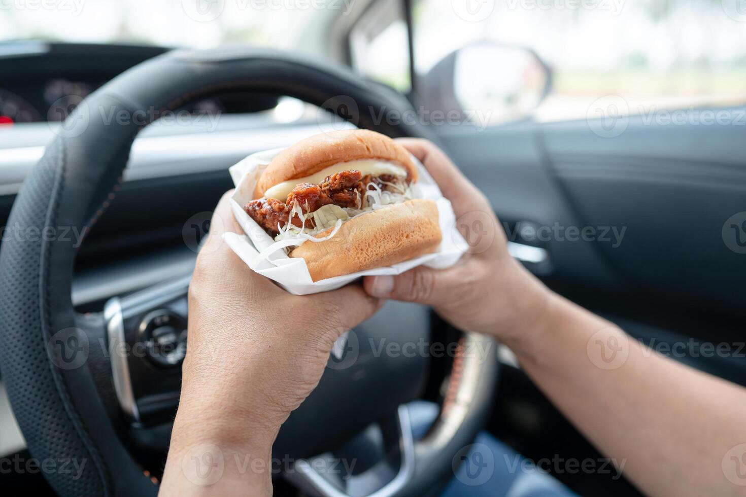 Asian lady holding hamburger to eat in car, dangerous and risk an accident. photo