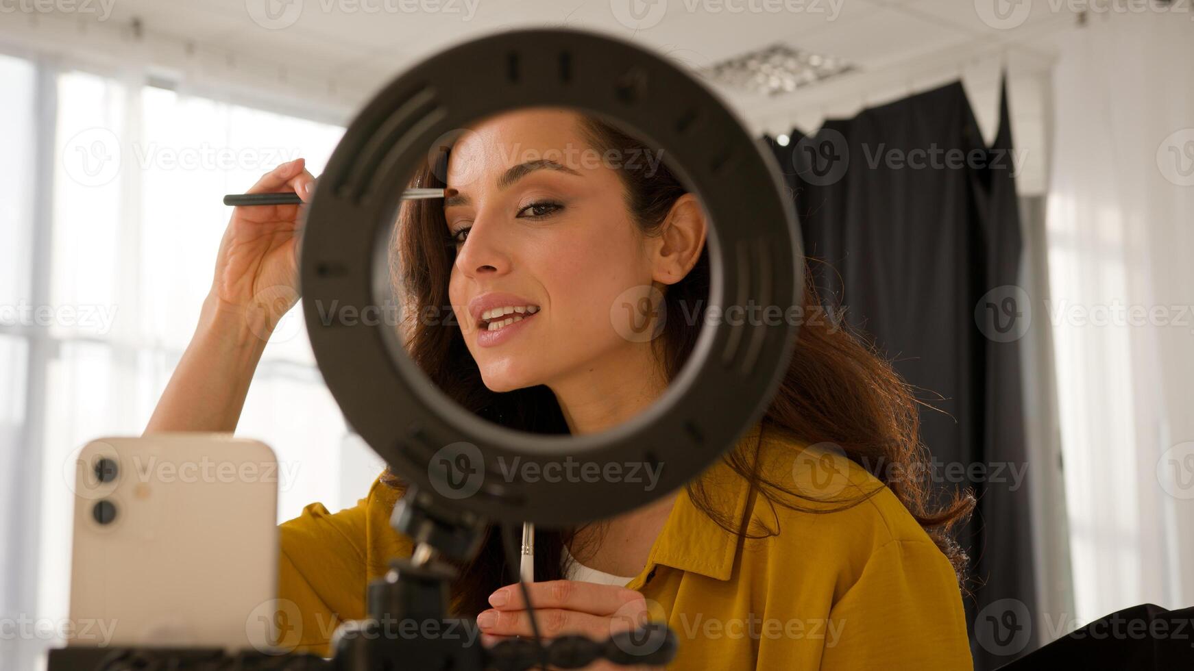 Glad Caucasian woman lady influencer trendy blogger recording vlog make-up blog streaming use mobile phone ring lamp stabilizer female girl vlogger apply eyeshadow cosmetics smile talk at home photo