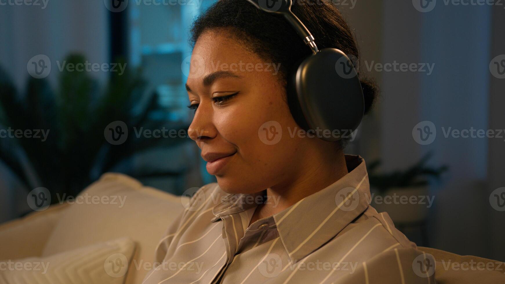Smiling African American woman in headphones on couch at evening listen song home leisure with modern tech female music lover listening mobile phone audio girl relaxing on couch in night living room photo
