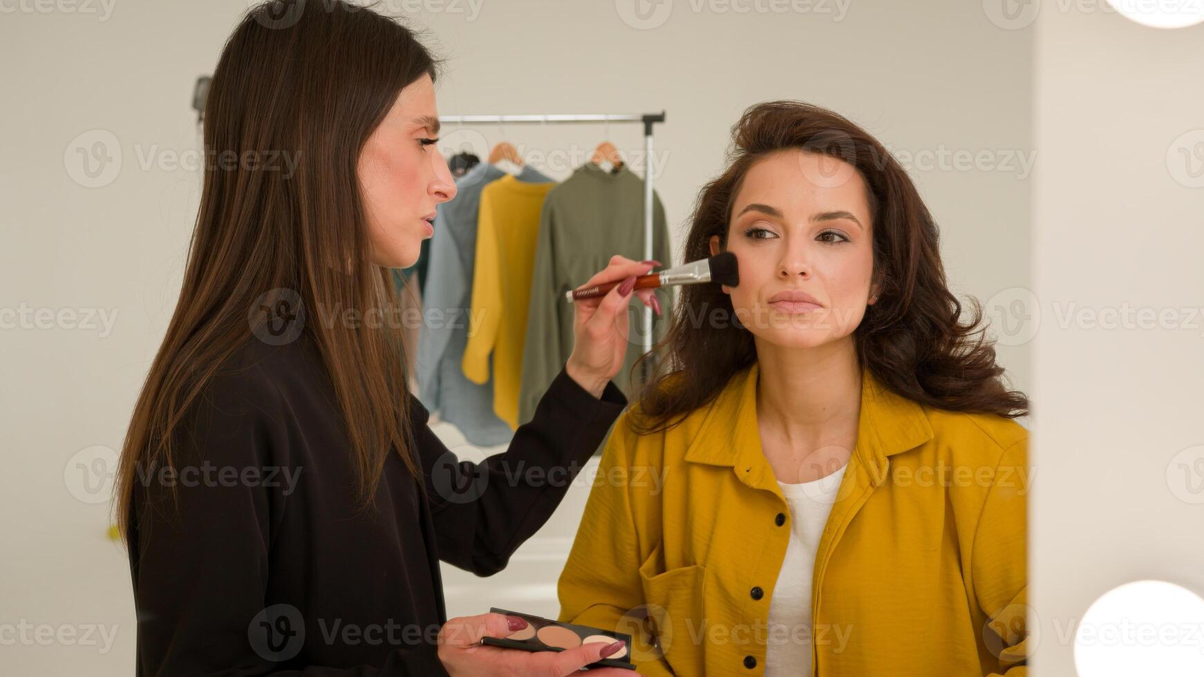 Caucasian woman makeup artist powdering girl trendy blogger in studio reflection female stylist applying shadows visage on influencer face brush getting ready for filming blog cosmetics fashion beauty photo