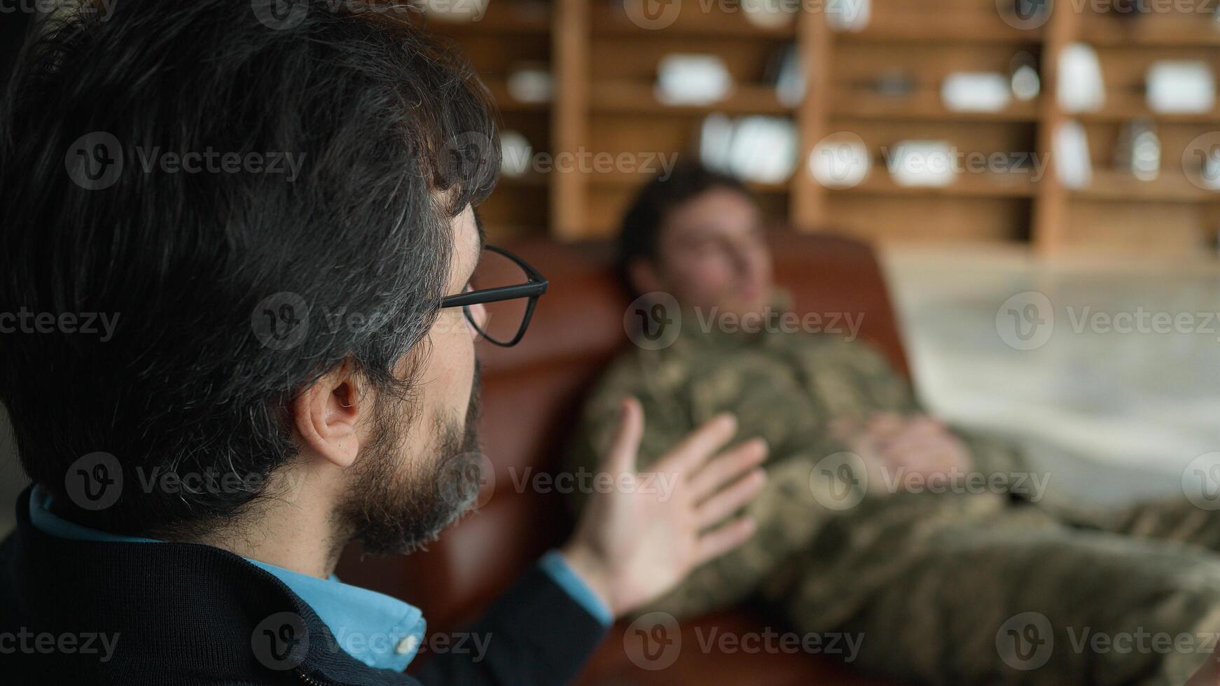 Middle aged Caucasian psychologist doctor man male speak talking to officer camouflage uniform military army soldier blurred patient guy lying on couch listening psychiatrist consultation session room photo