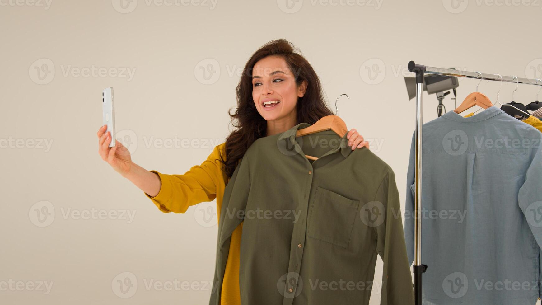 Carefree Caucasian blogger girl streamer smile speaking filming blog influencer use mobile phone in studio. Glad woman model compare clothes fashion recording vlog on smartphone female streaming photo