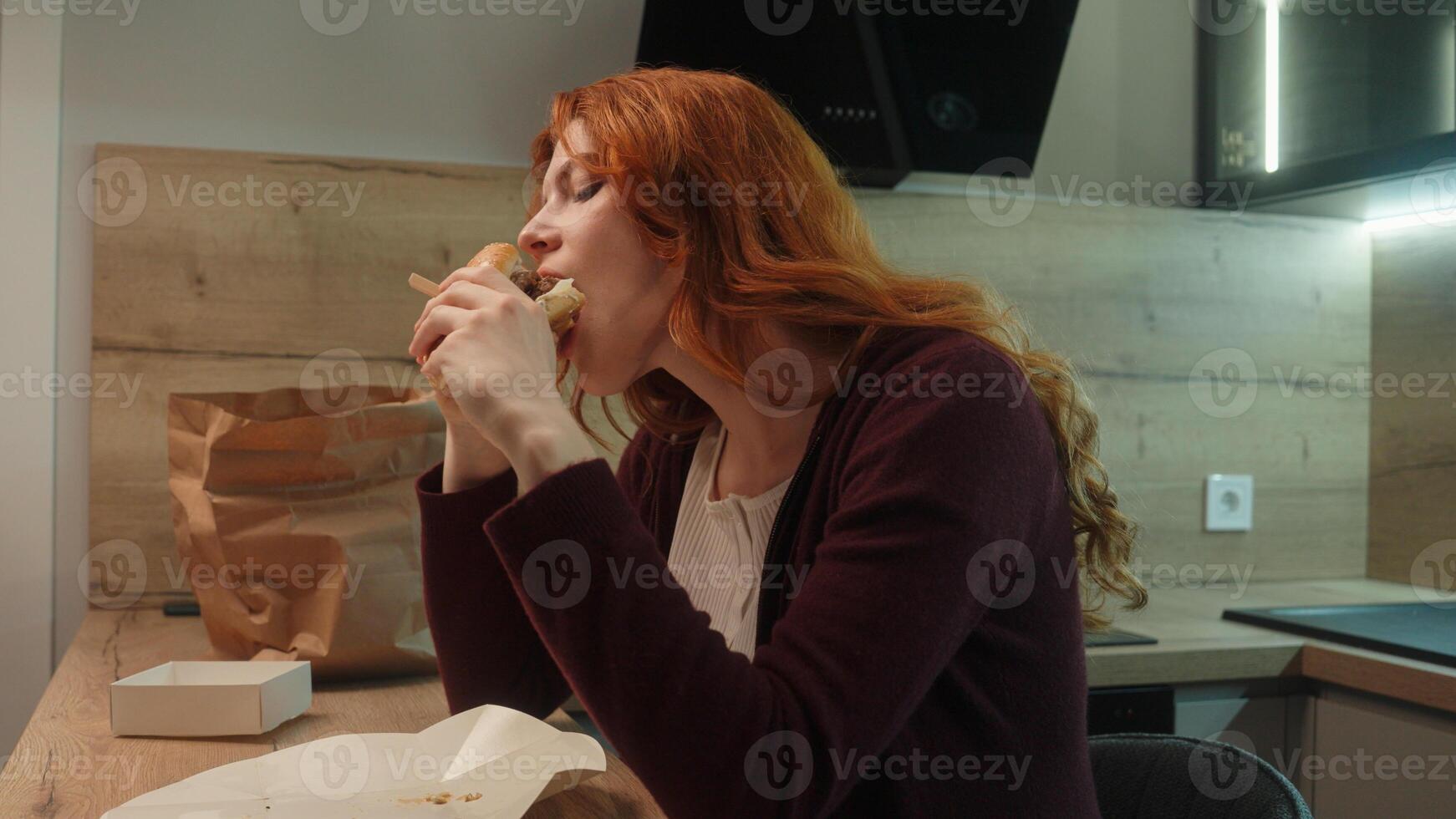 Young Caucasian happy client woman buyer female girl satisfied shopper customer enjoy chewing bite fast food tasting eat tasty burger cheeseburger at kitchen overweight unhealthy eating calorie weight photo