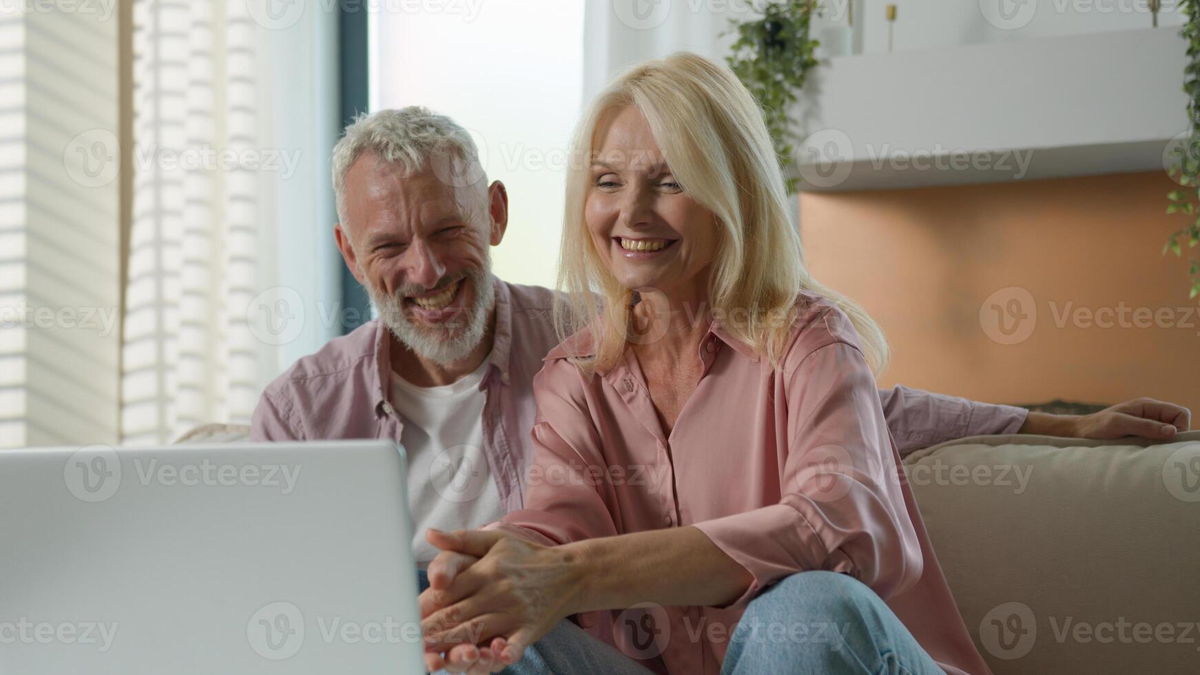 Mature senior middle aged Caucasian couple smiling cheerful woman man laugh together old family looking screen computer laptop pc talking at home sofa female male relaxing happy relationship good mood photo