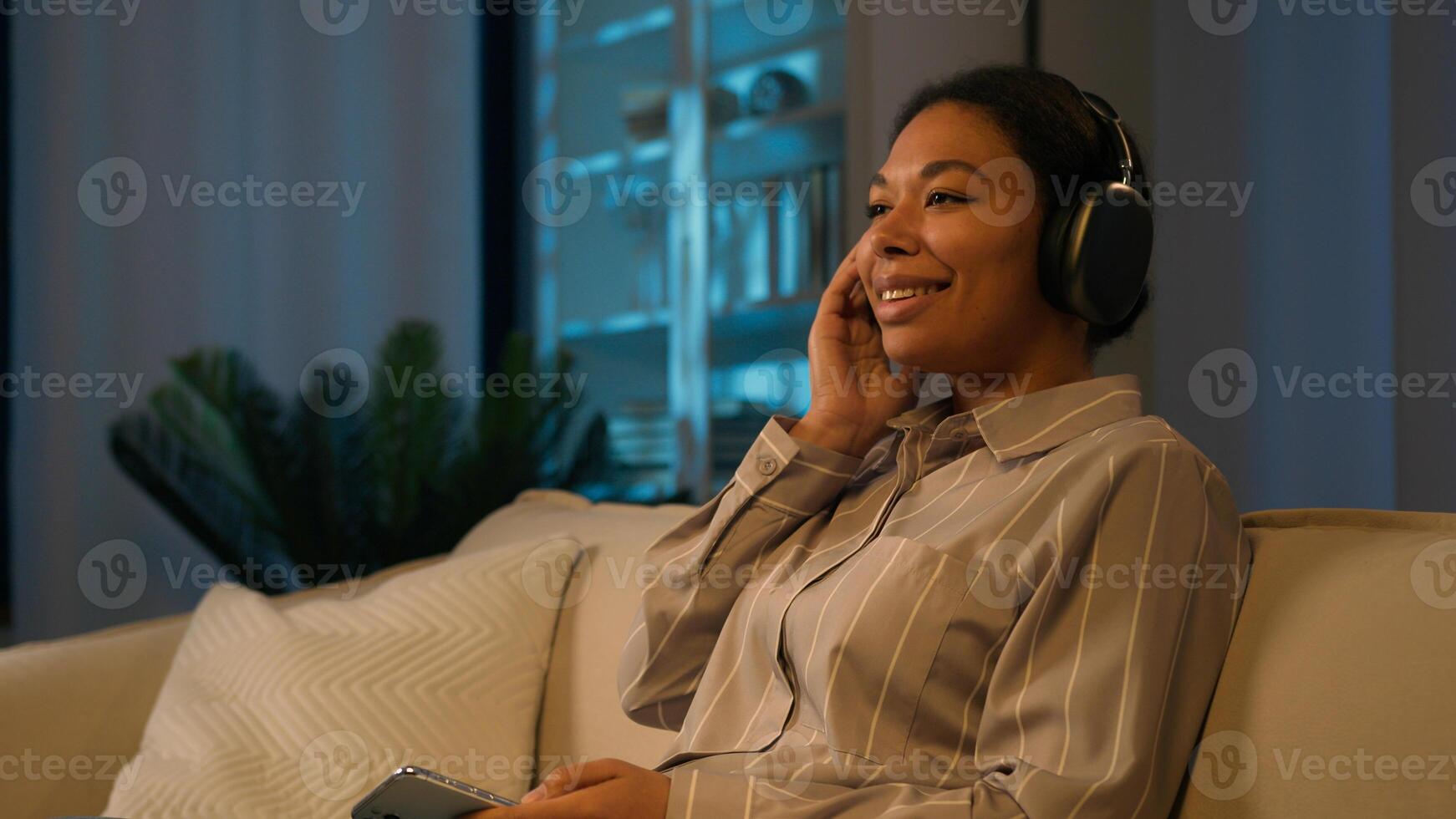 Smiling African American woman in headphones on couch at evening listen song home leisure with modern tech female music lover listening mobile phone audio girl relaxing on couch in night living room photo