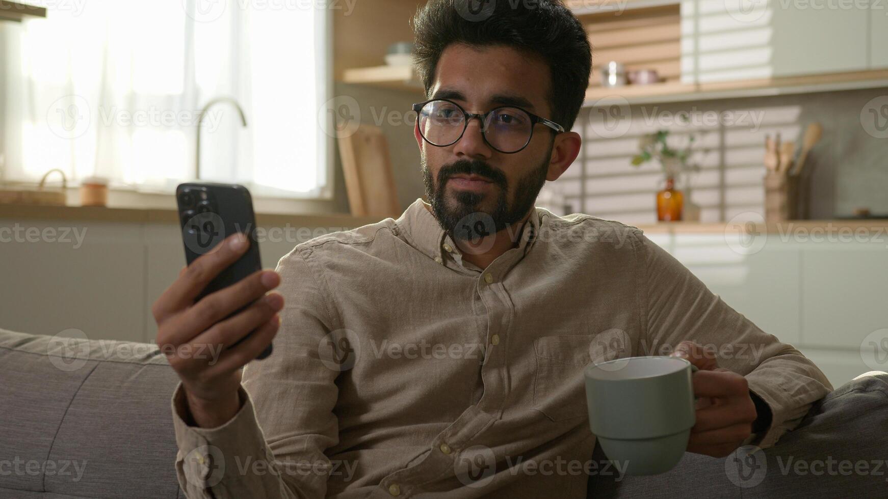 Relaxing Arabian man male ethnic guy on comfortable couch sofa using mobile phone drink coffee at kitchen Indian millennial freelancer working from home read news on smartphone looking at camera smile photo