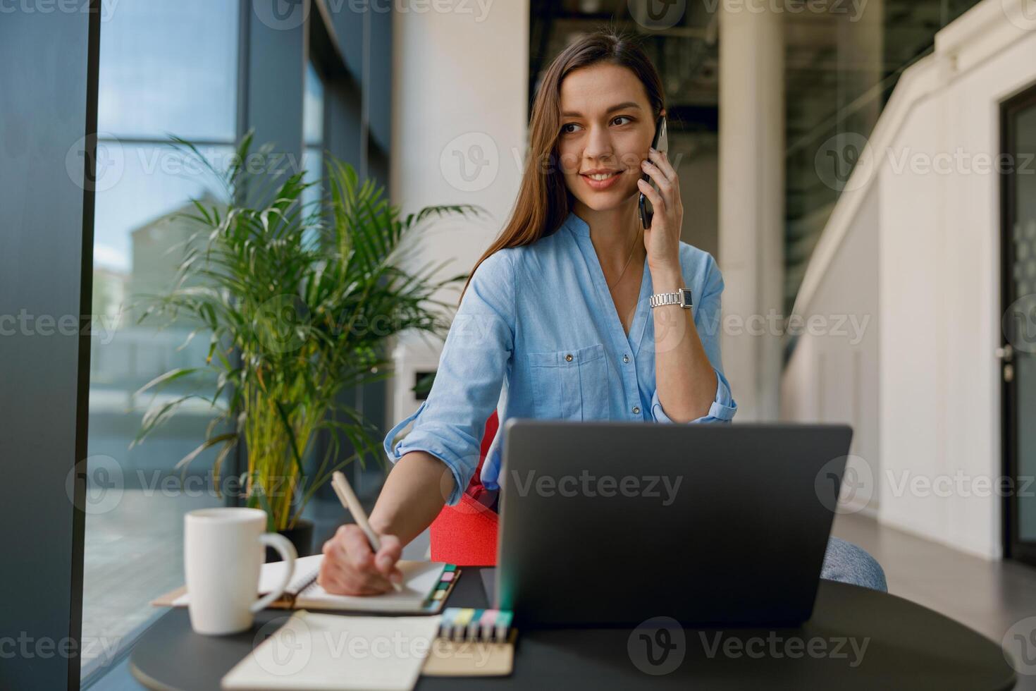 A businesswoman is in her professional workspace, multitasking by taking notes on a call photo