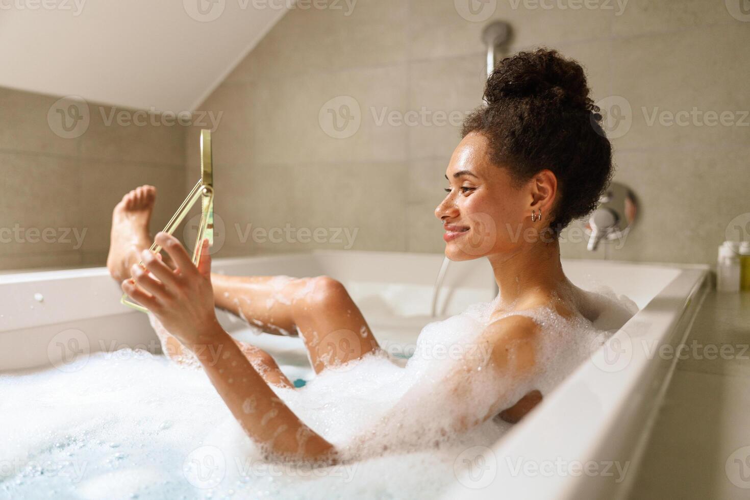 Woman in bathtub, phone in hand, happy expression, shoulder and chest visible photo