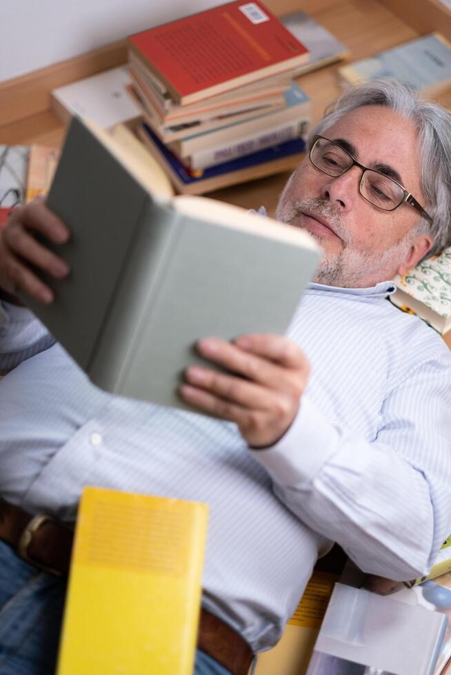 Good-looking 60-year-old man engages in reading an interesting book lying on the floor among many books. photo