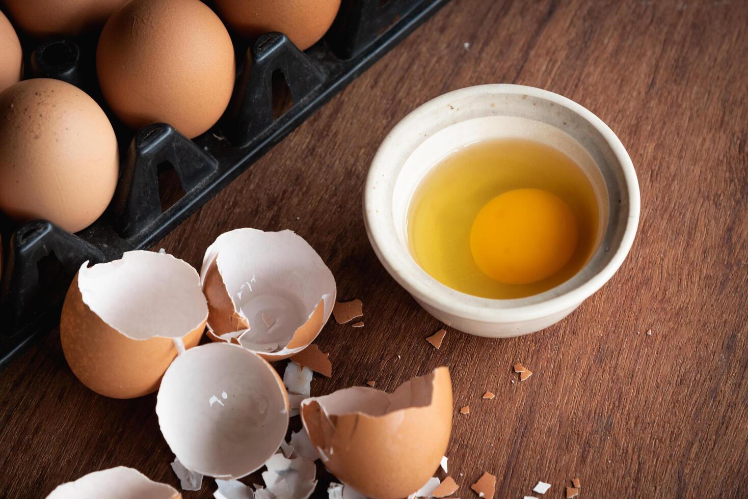 Broken egg shells and eggs on tray and pot, raw egg in small bowl at wooden background menu and recipe photo