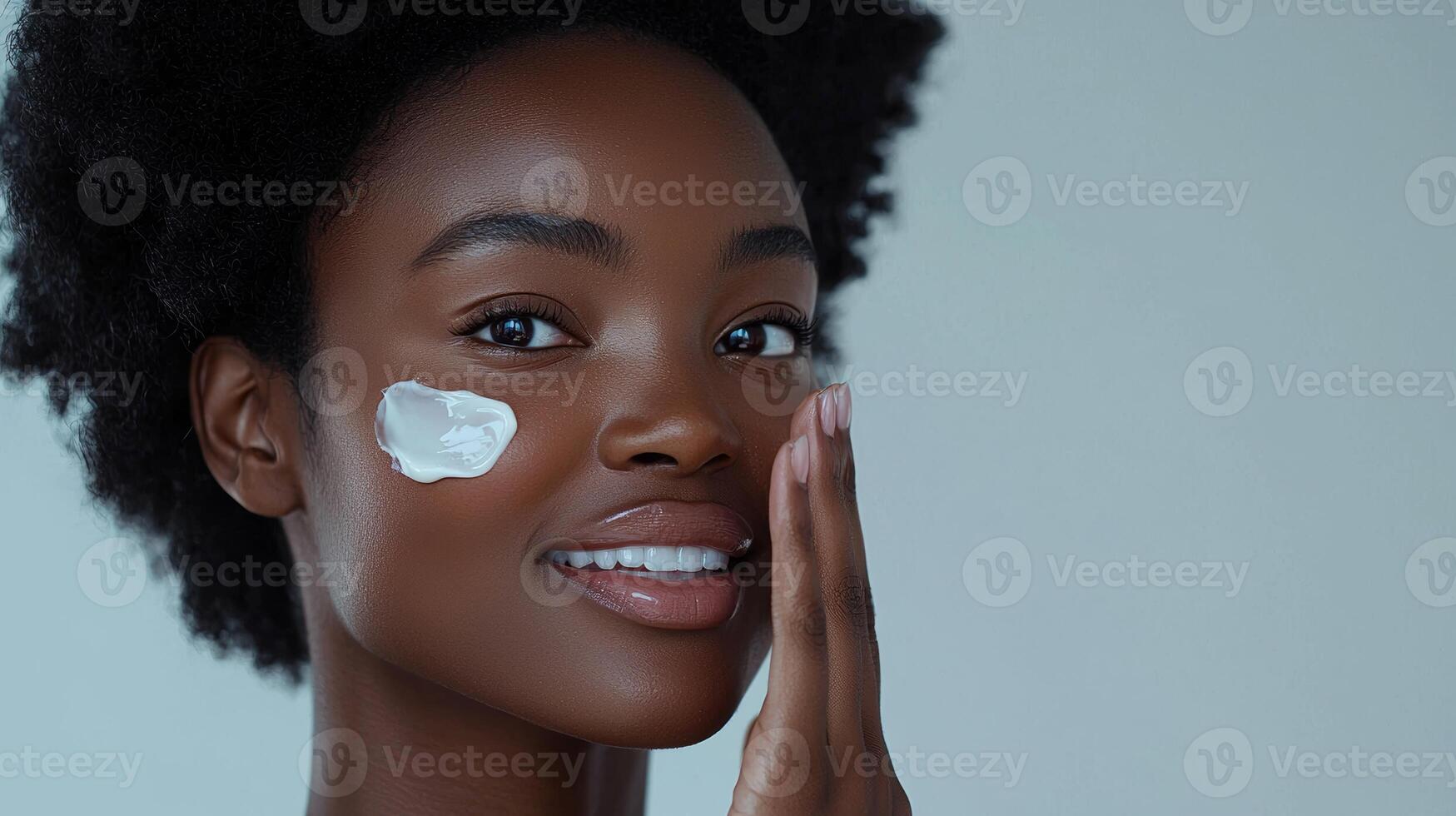 Advertisement image of a Black woman with flawless skin, softly smiling as she applies a moisturizing cream to her cheek with her fingertips, set against a clean, neutral gray background, perfect photo