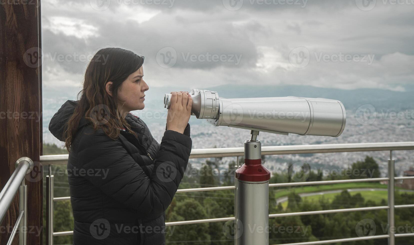 woman, standing at a viewing platform, holding in her hands long binoculars mounted on a metal pedestal against a cloudy sky background photo
