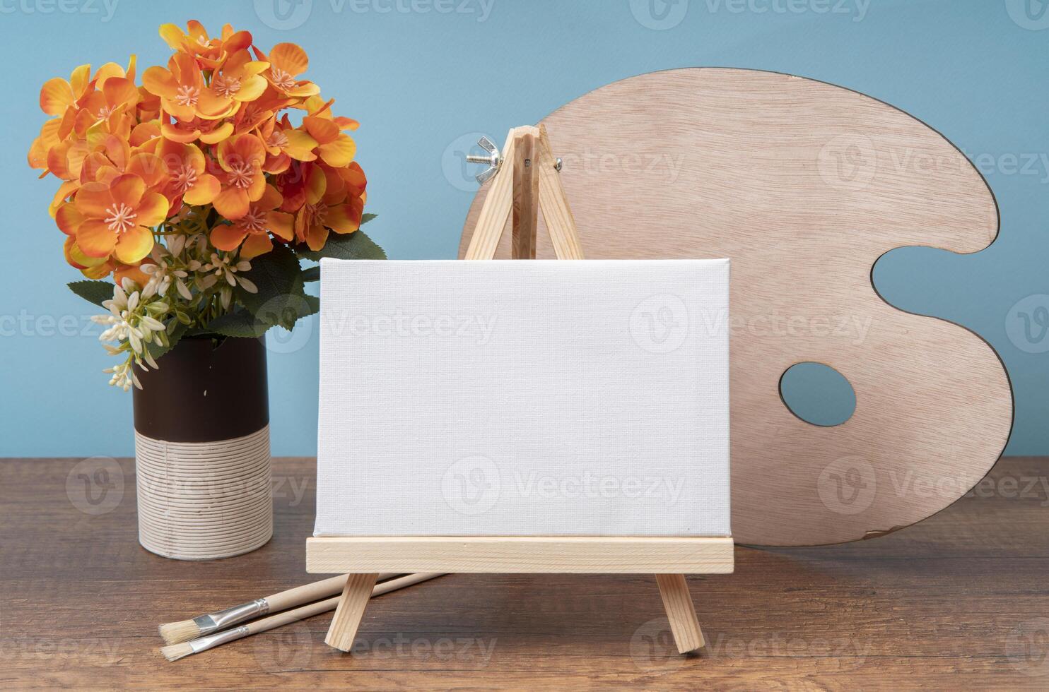 small wooden easel with blank canvas on wooden table decorated with glass with flowers, paintbrushes and palette photo