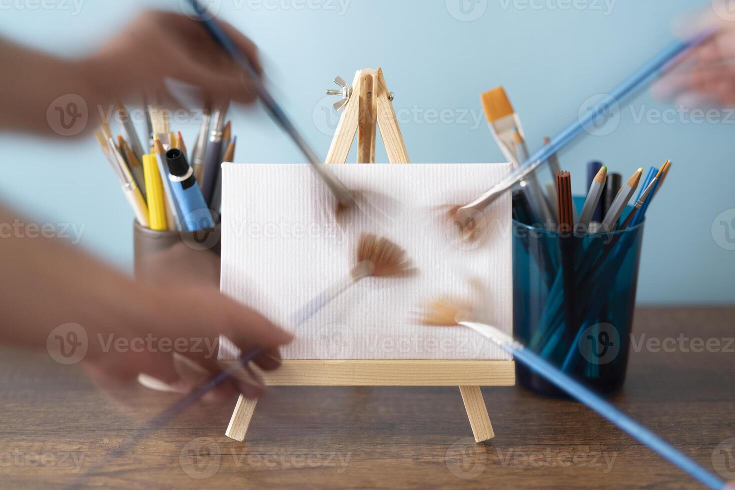 moving paintbrush making brush strokes on small wooden easel with blank canvas on wooden table decorated with glass with paint tubes, brushes and pencils photo