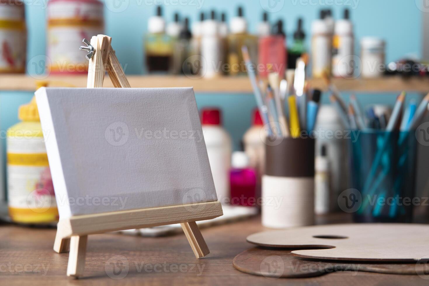 small wooden easel with blank canvas on wooden table against background of shelves with paint tubes, brushes and pencils photo