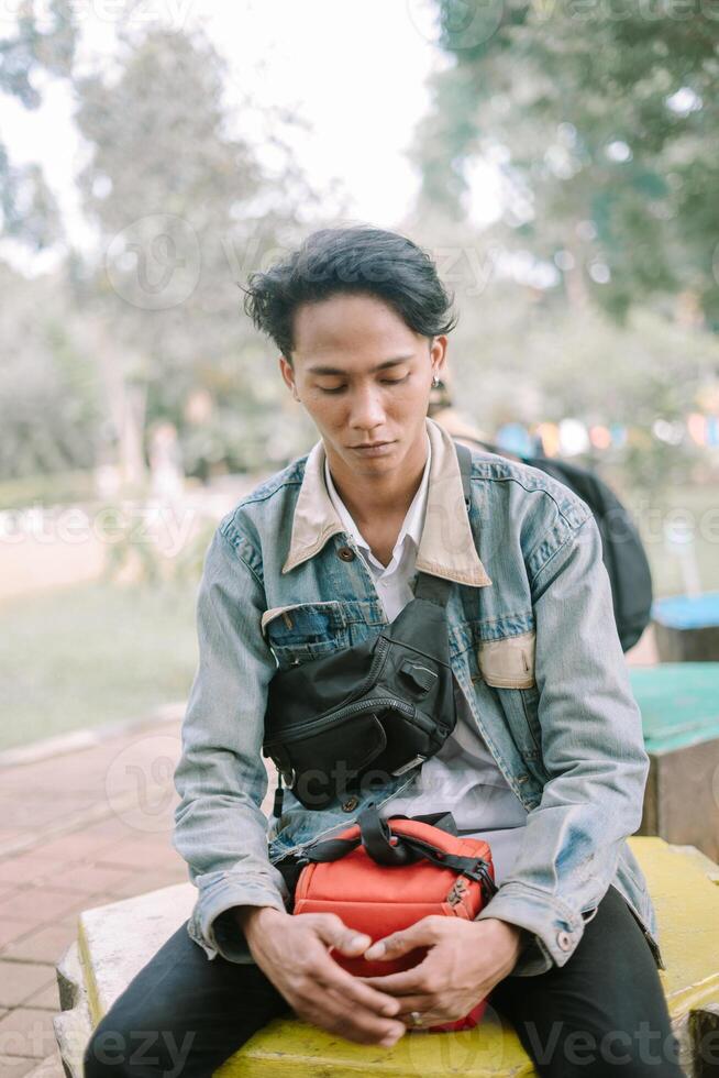 A Southeast Asian man wearing a denim jacket, sitting on a bench in a city square, photo