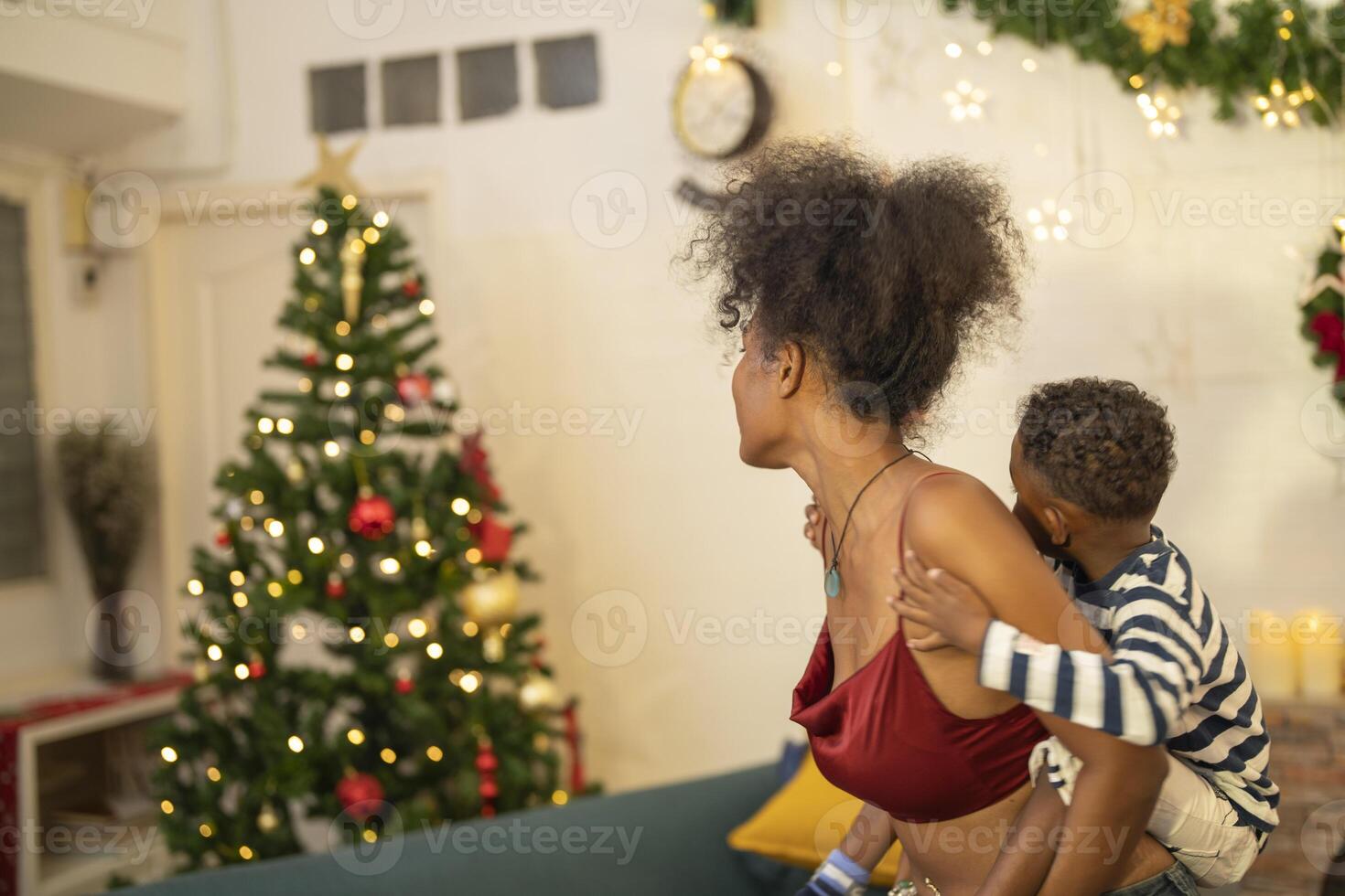 mother in a festive setting with a beautifully Christmas tree engages playfully with her child, who is lying on couch. The joyful atmosphere highlights holiday family bonding, home celebrations. photo