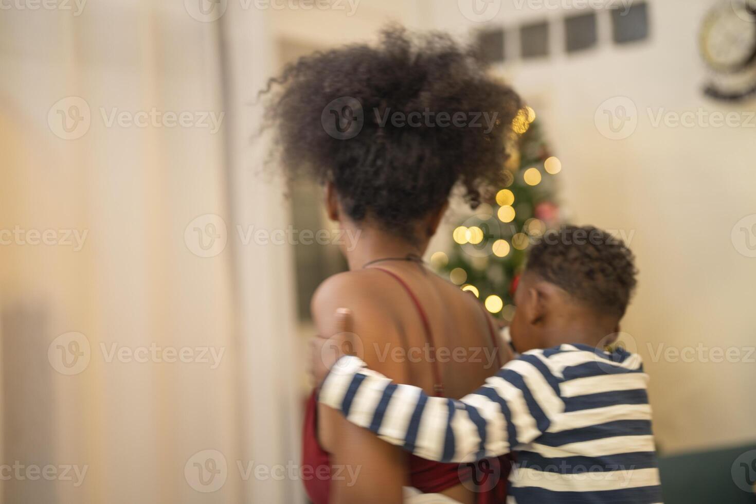 mother in a festive setting with a beautifully Christmas tree engages playfully with her child, who is lying on couch. The joyful atmosphere highlights holiday family bonding, home celebrations. photo