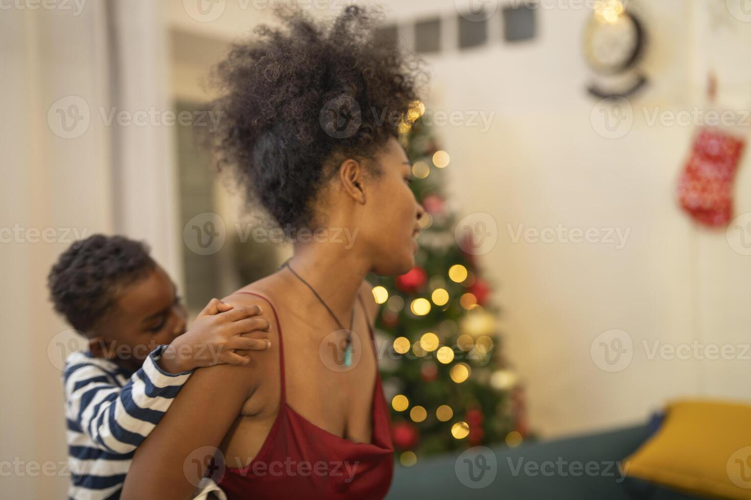 mother in a festive setting with a beautifully Christmas tree engages playfully with her child, who is lying on couch. The joyful atmosphere highlights holiday family bonding, home celebrations. photo