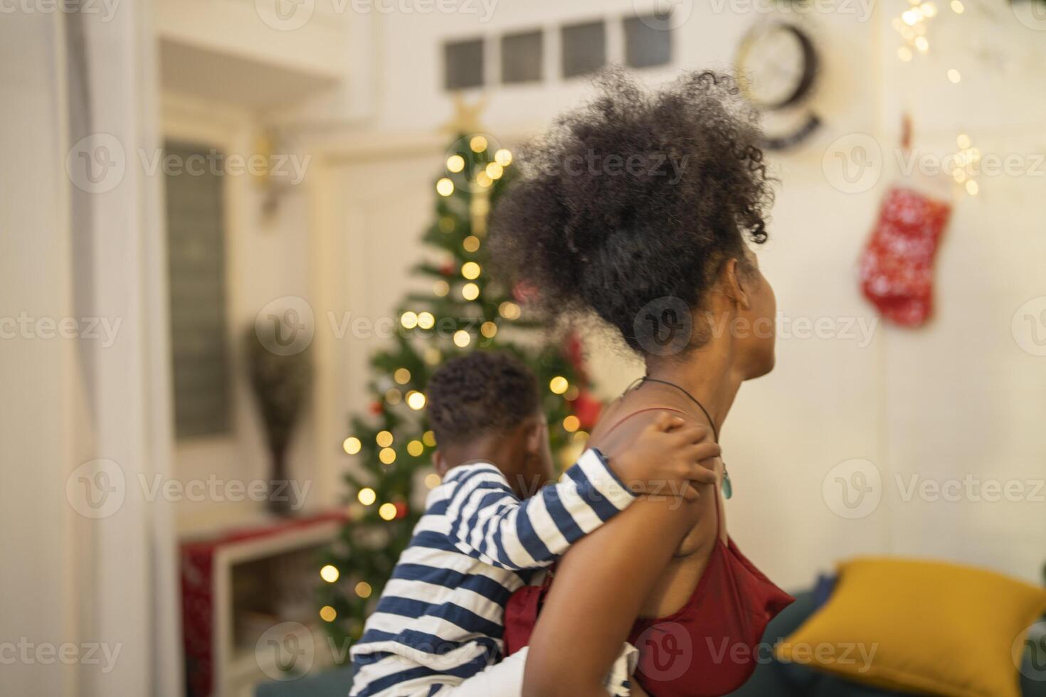 mother in a festive setting with a beautifully Christmas tree engages playfully with her child, who is lying on couch. The joyful atmosphere highlights holiday family bonding, home celebrations. photo