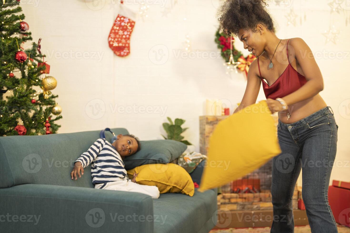 mother in a festive setting with a beautifully Christmas tree engages playfully with her child, who is lying on couch. The joyful atmosphere highlights holiday family bonding, home celebrations. photo