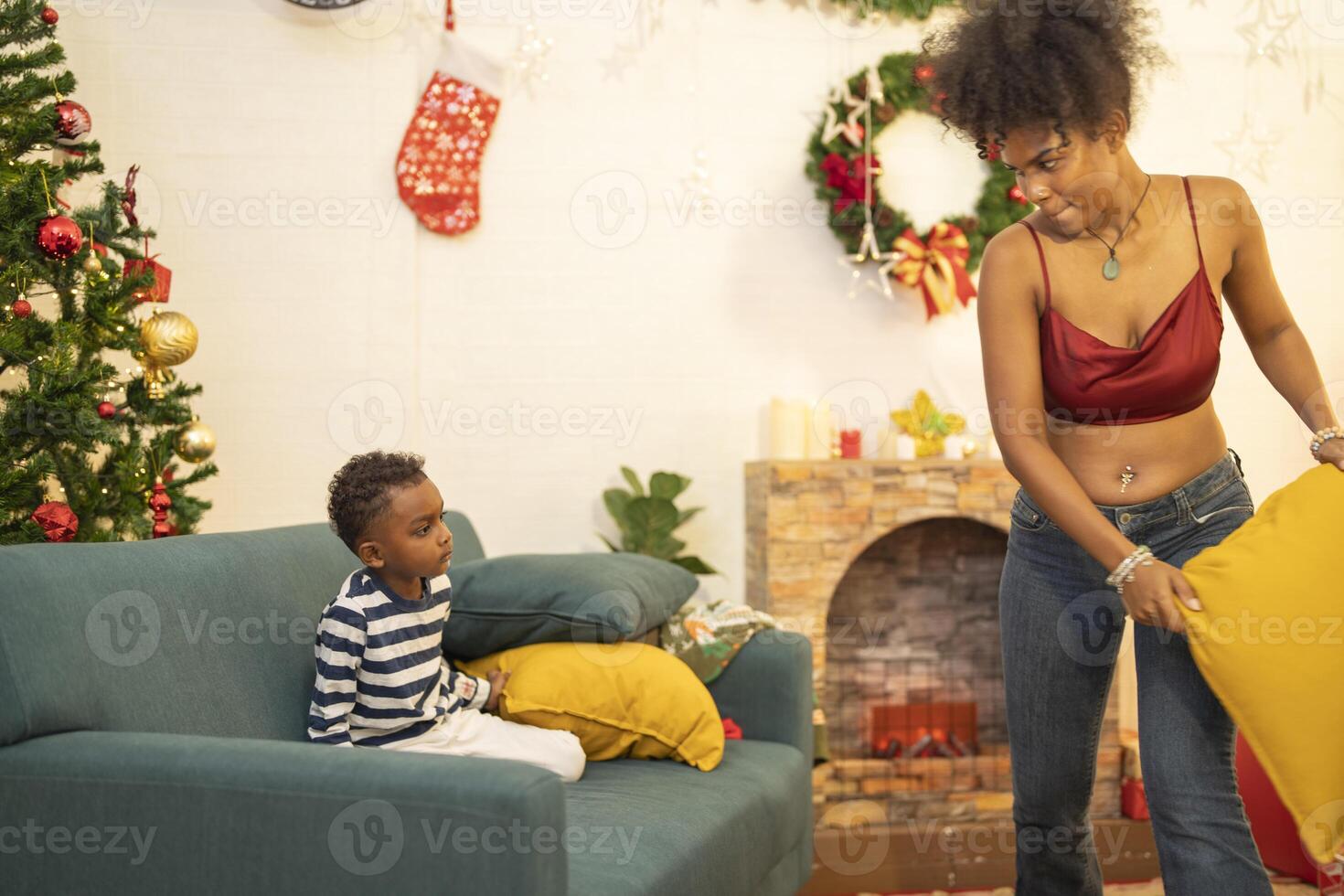 mother in a festive setting with a beautifully Christmas tree engages playfully with her child, who is lying on couch. The joyful atmosphere highlights holiday family bonding, home celebrations. photo