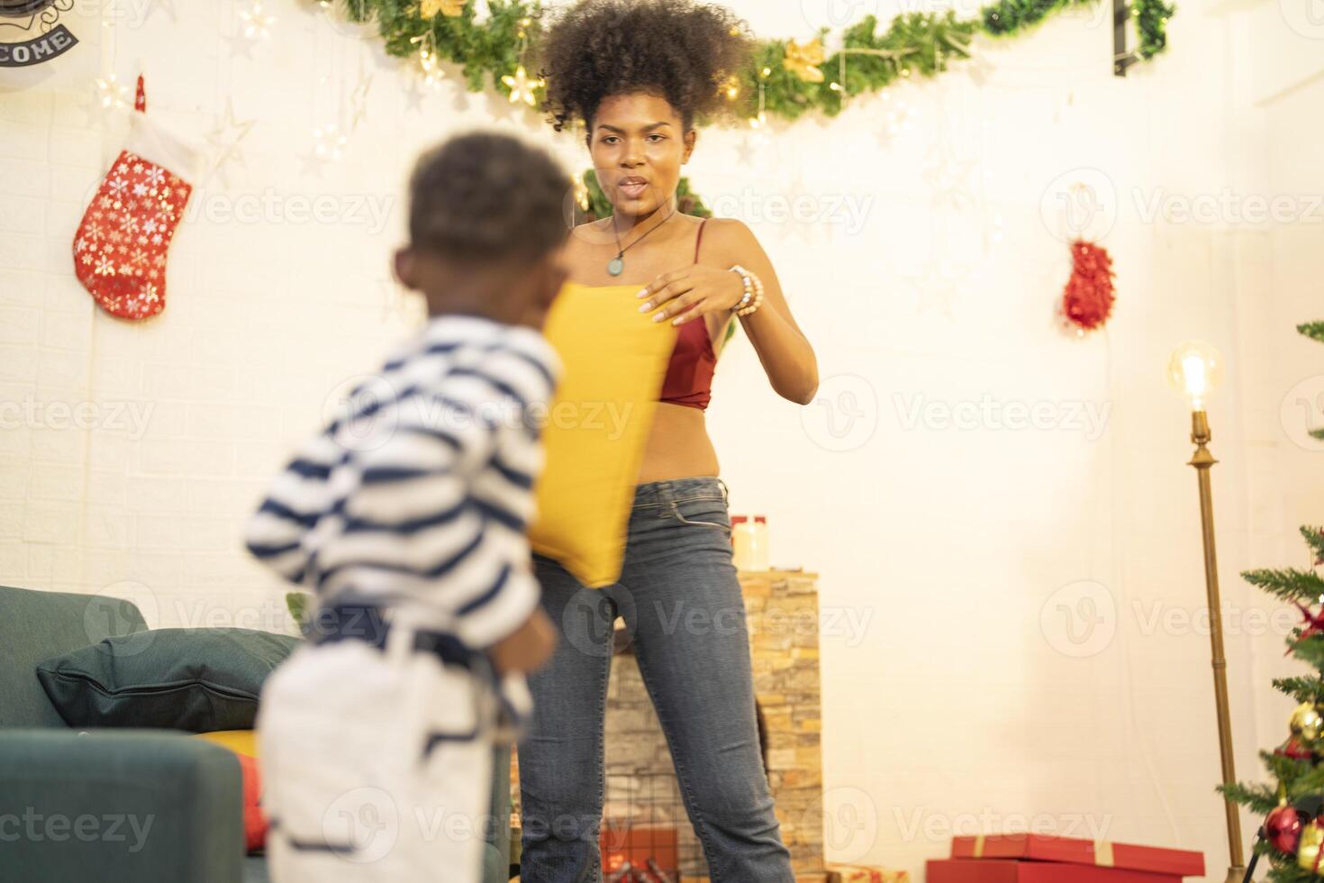 mother in a festive setting with a beautifully Christmas tree engages playfully with her child, who is lying on couch. The joyful atmosphere highlights holiday family bonding, home celebrations. photo