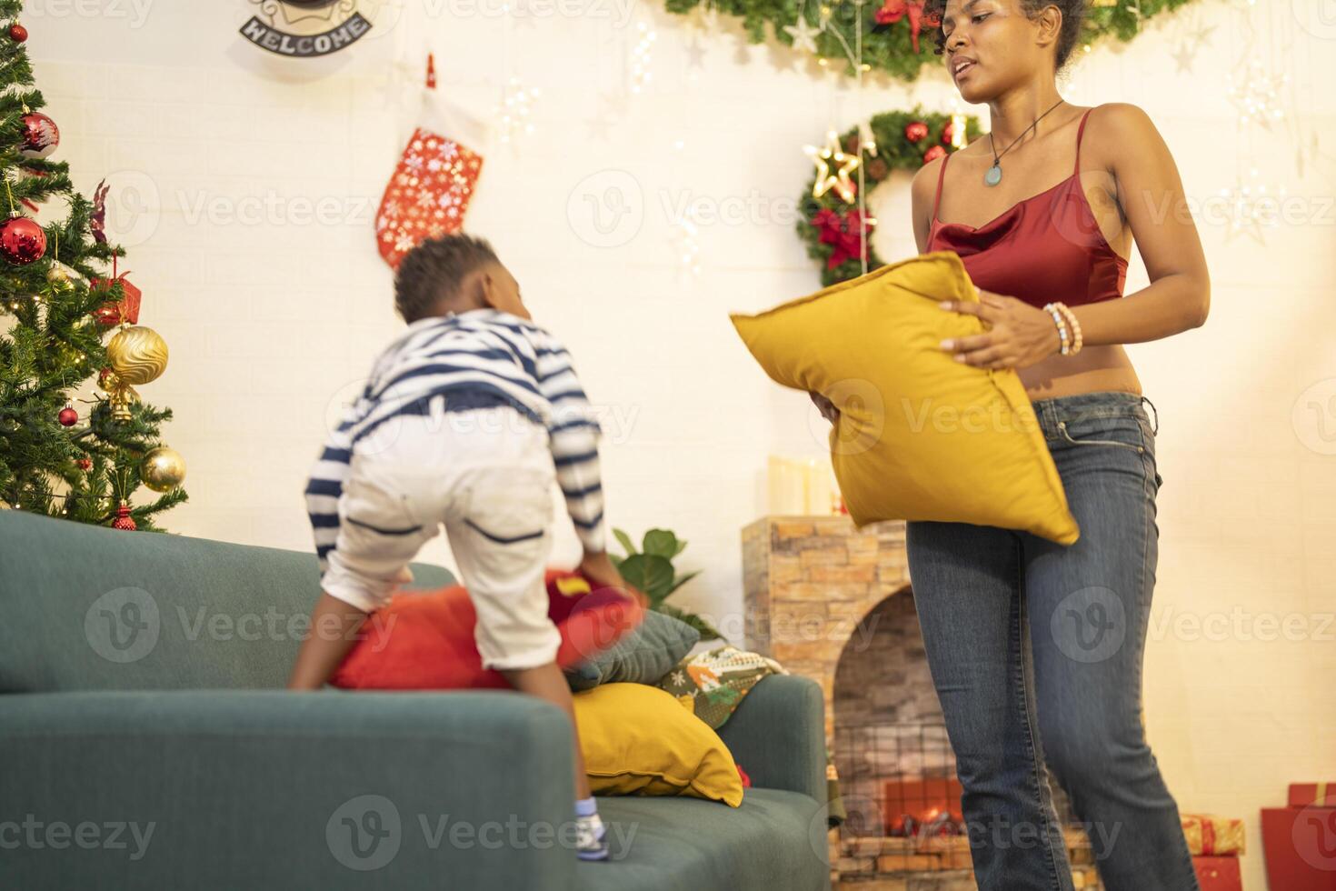 mother in a festive setting with a beautifully Christmas tree engages playfully with her child, who is lying on couch. The joyful atmosphere highlights holiday family bonding, home celebrations. photo