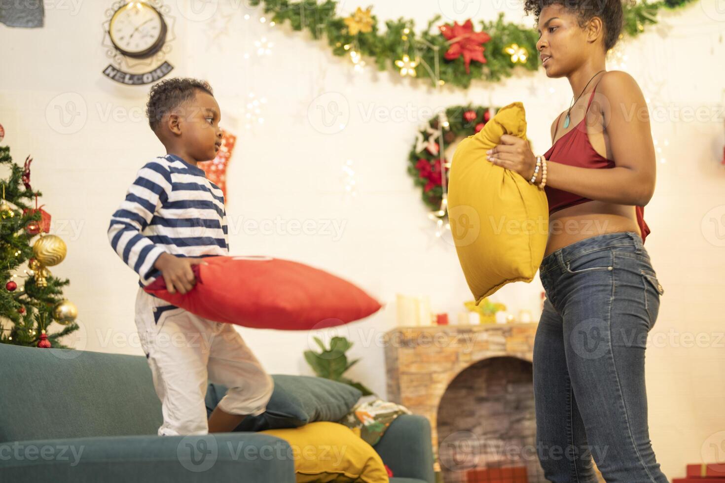 mother in a festive setting with a beautifully Christmas tree engages playfully with her child, who is lying on couch. The joyful atmosphere highlights holiday family bonding, home celebrations. photo
