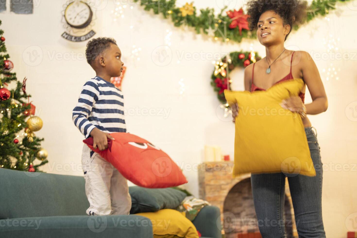 mother in a festive setting with a beautifully Christmas tree engages playfully with her child, who is lying on couch. The joyful atmosphere highlights holiday family bonding, home celebrations. photo