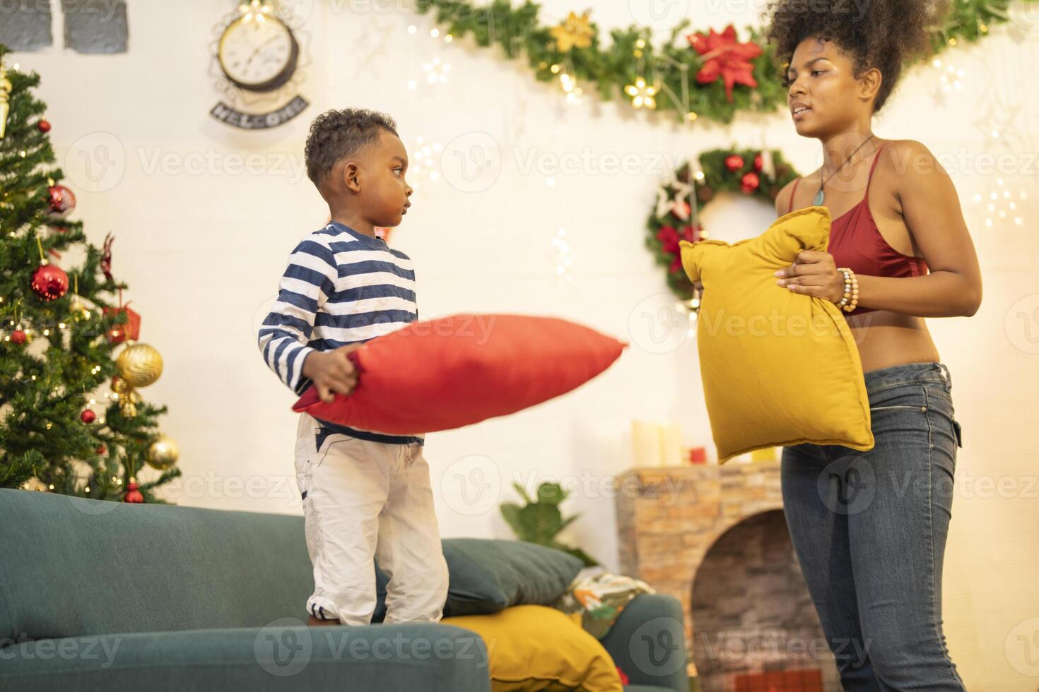 mother in a festive setting with a beautifully Christmas tree engages playfully with her child, who is lying on couch. The joyful atmosphere highlights holiday family bonding, home celebrations. photo