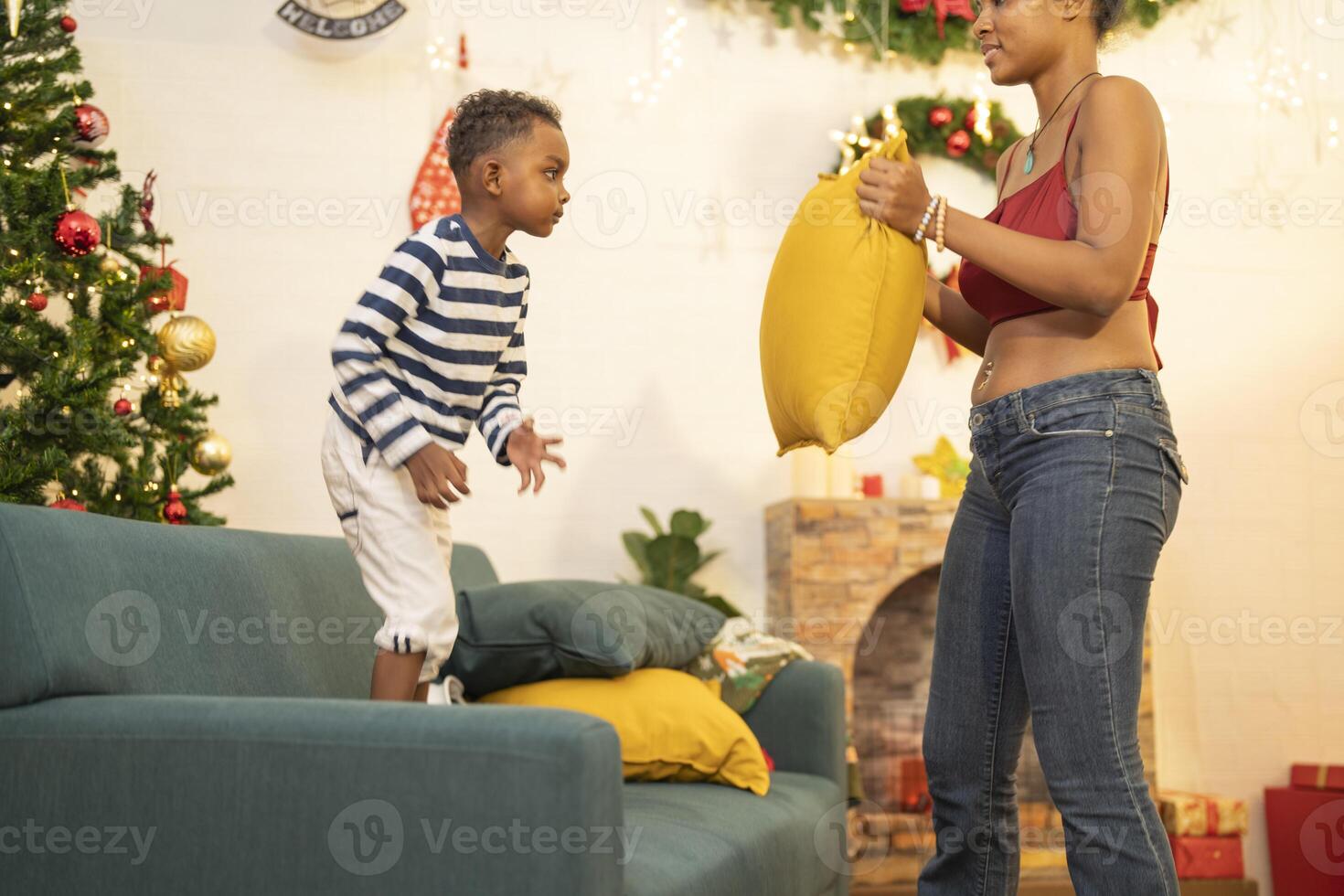 mother in a festive setting with a beautifully Christmas tree engages playfully with her child, who is lying on couch. The joyful atmosphere highlights holiday family bonding, home celebrations. photo