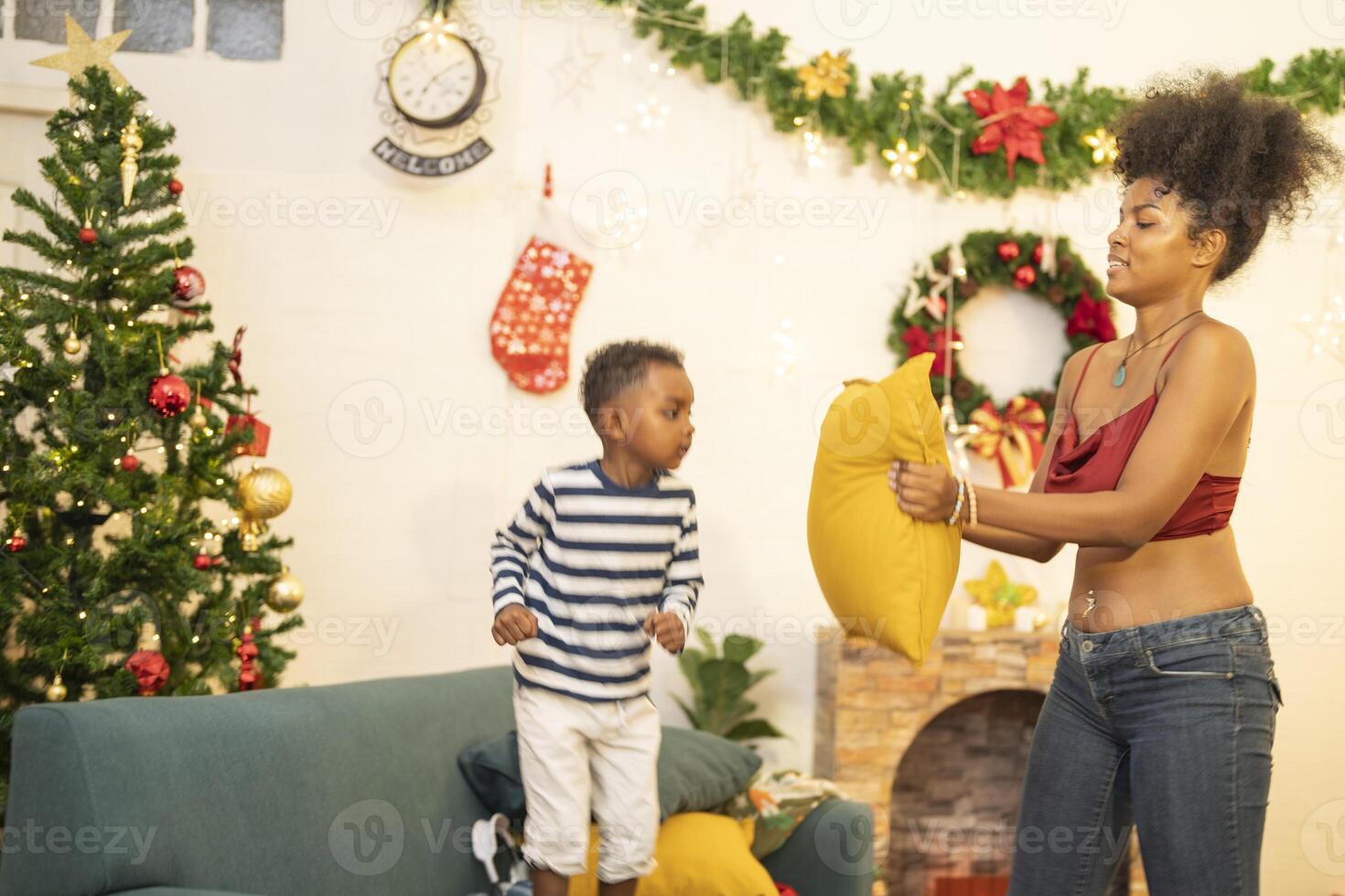 mother in a festive setting with a beautifully Christmas tree engages playfully with her child, who is lying on couch. The joyful atmosphere highlights holiday family bonding, home celebrations. photo