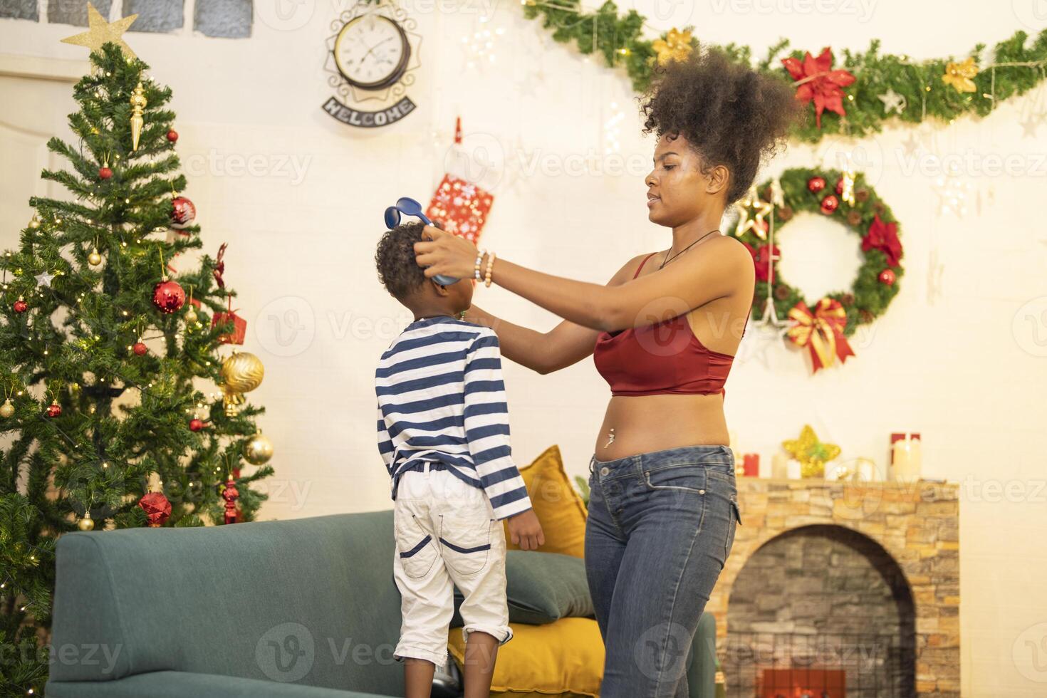 mother in a festive setting with a beautifully Christmas tree engages playfully with her child, who is lying on couch. The joyful atmosphere highlights holiday family bonding, home celebrations. photo