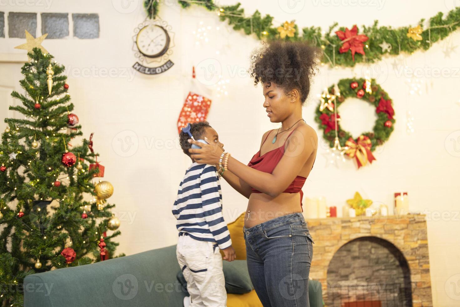 mother in a festive setting with a beautifully Christmas tree engages playfully with her child, who is lying on couch. The joyful atmosphere highlights holiday family bonding, home celebrations. photo