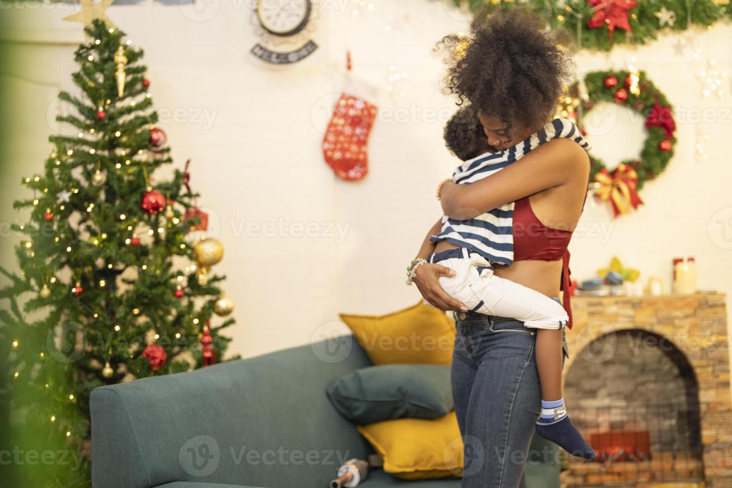 mother in a festive setting with a beautifully Christmas tree engages playfully with her child, who is lying on couch. The joyful atmosphere highlights holiday family bonding, home celebrations. photo