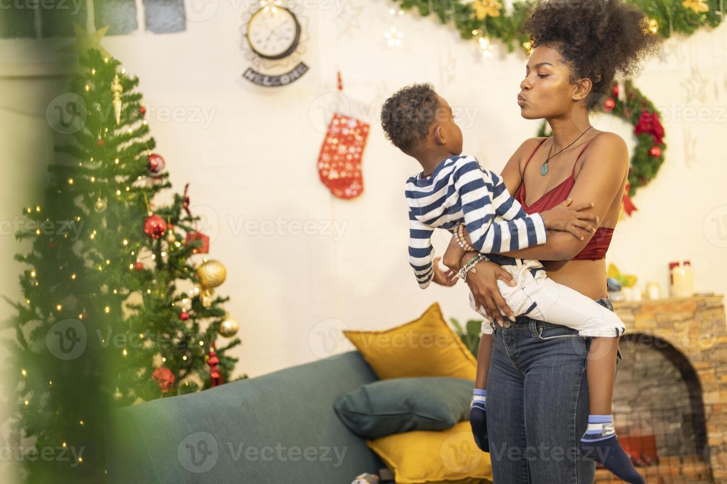mother in a festive setting with a beautifully Christmas tree engages playfully with her child, who is lying on couch. The joyful atmosphere highlights holiday family bonding, home celebrations. photo