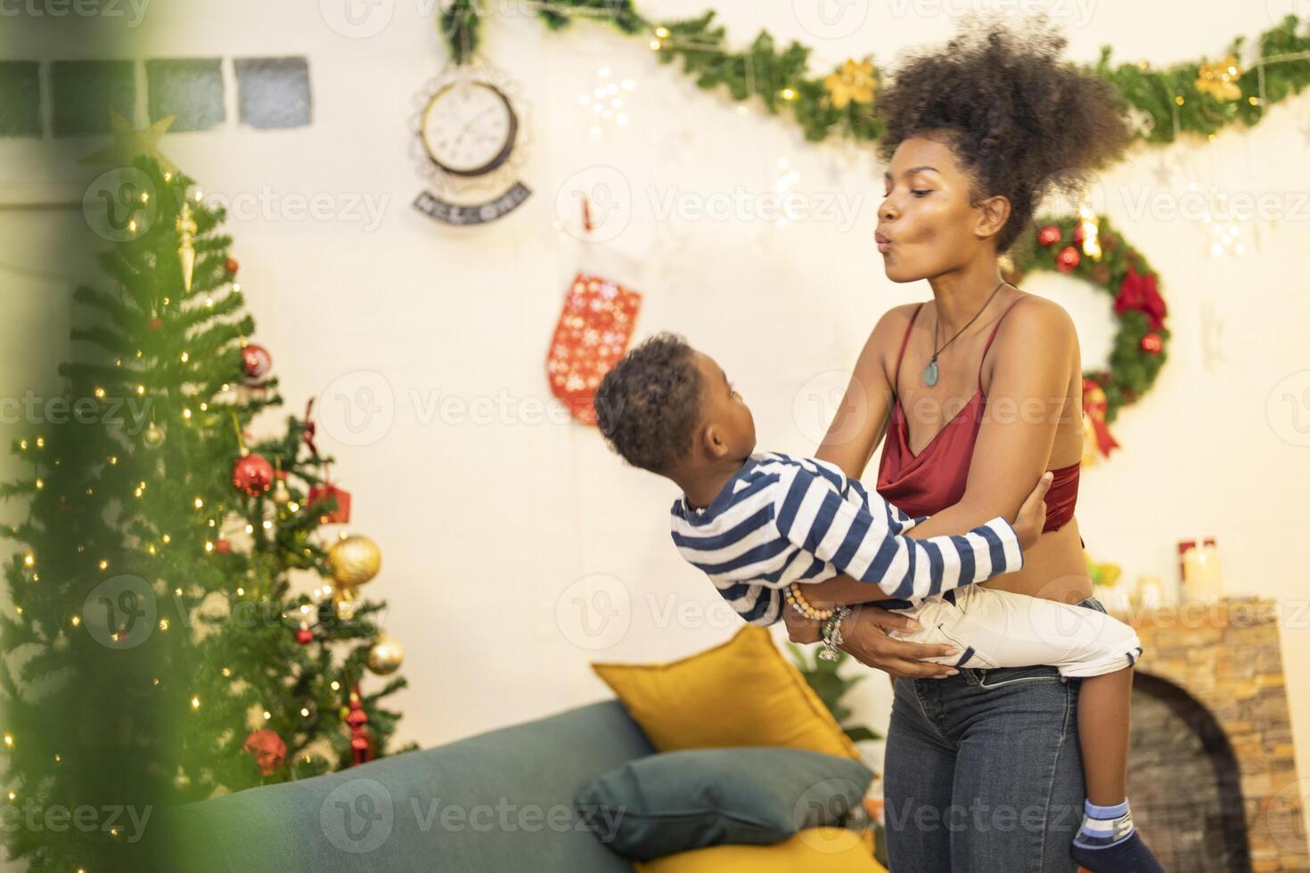 mother in a festive setting with a beautifully Christmas tree engages playfully with her child, who is lying on couch. The joyful atmosphere highlights holiday family bonding, home celebrations. photo