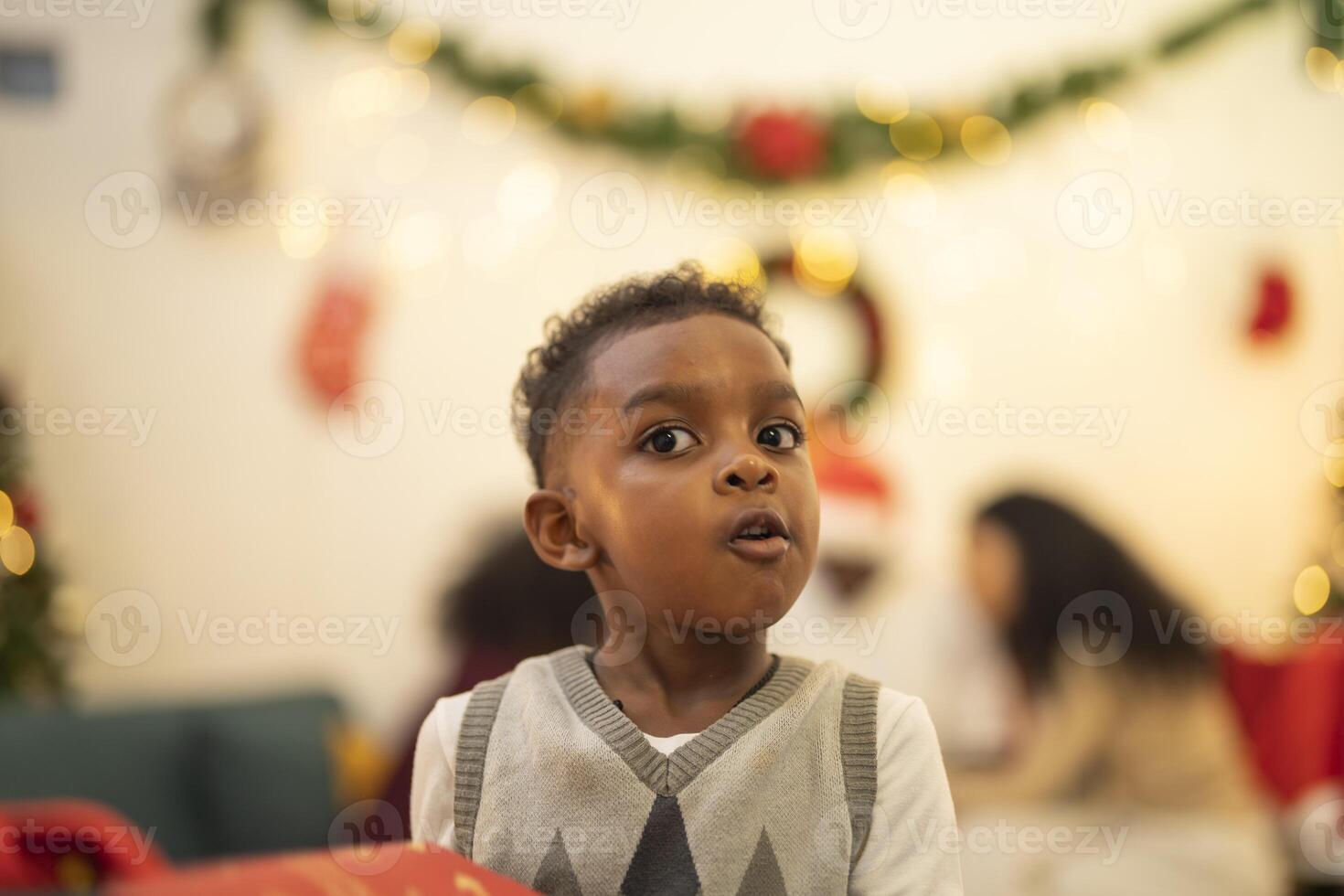 A young child excitedly holding a Christmas gift box, surrounded by festive decorations and twinkling lights in the background. The joyful expression on the child's face reflects the happiness. photo
