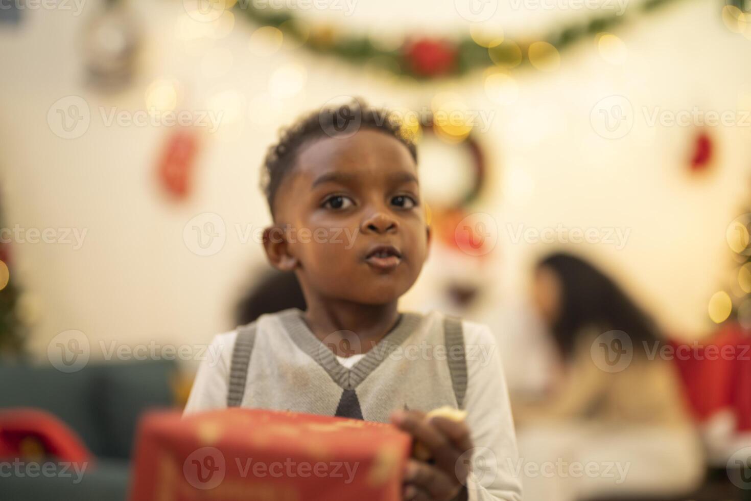 A young child excitedly holding a Christmas gift box, surrounded by festive decorations and twinkling lights in the background. The joyful expression on the child's face reflects the happiness. photo