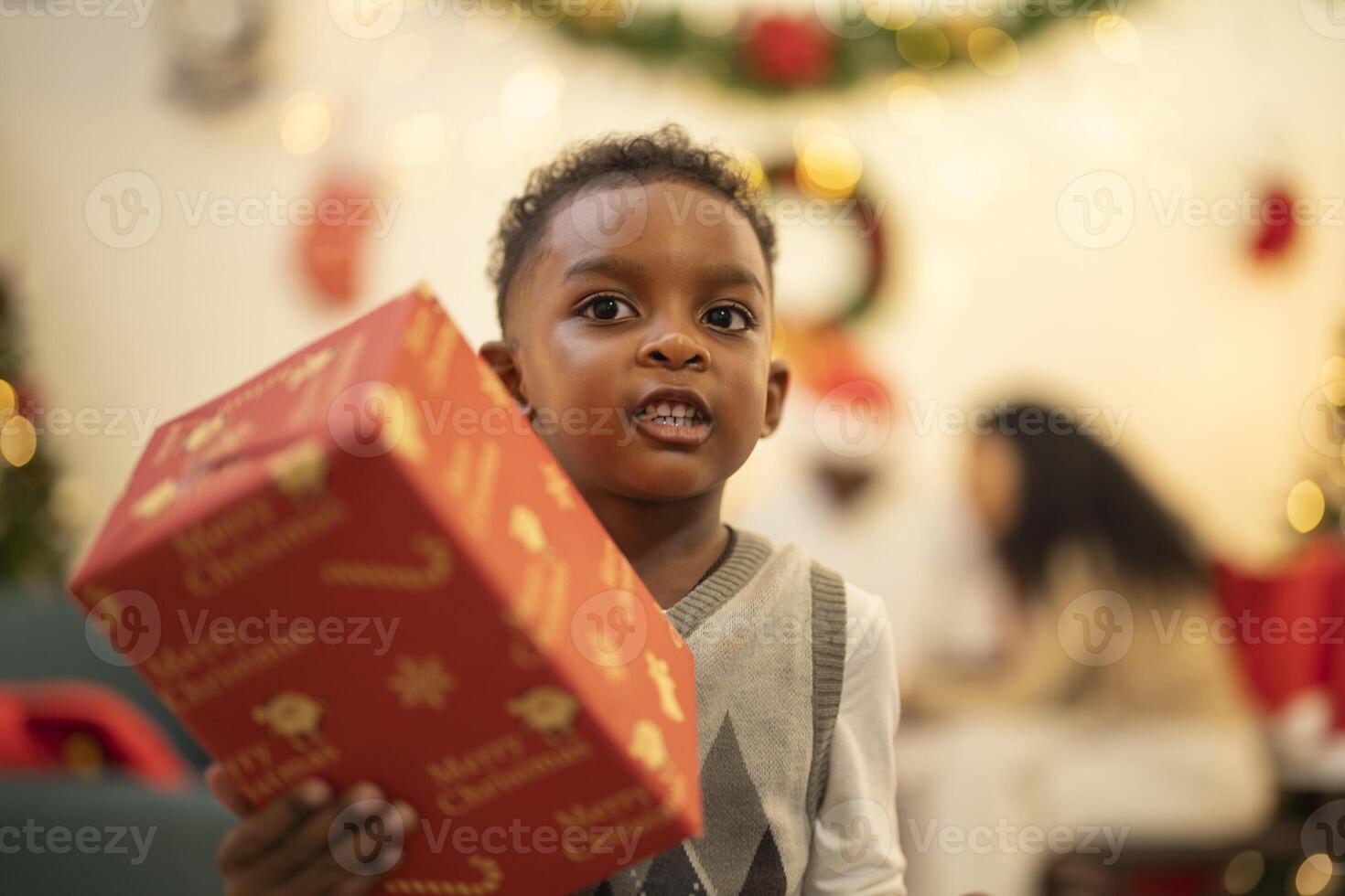 A young child excitedly holding a Christmas gift box, surrounded by festive decorations and twinkling lights in the background. The joyful expression on the child's face reflects the happiness. photo