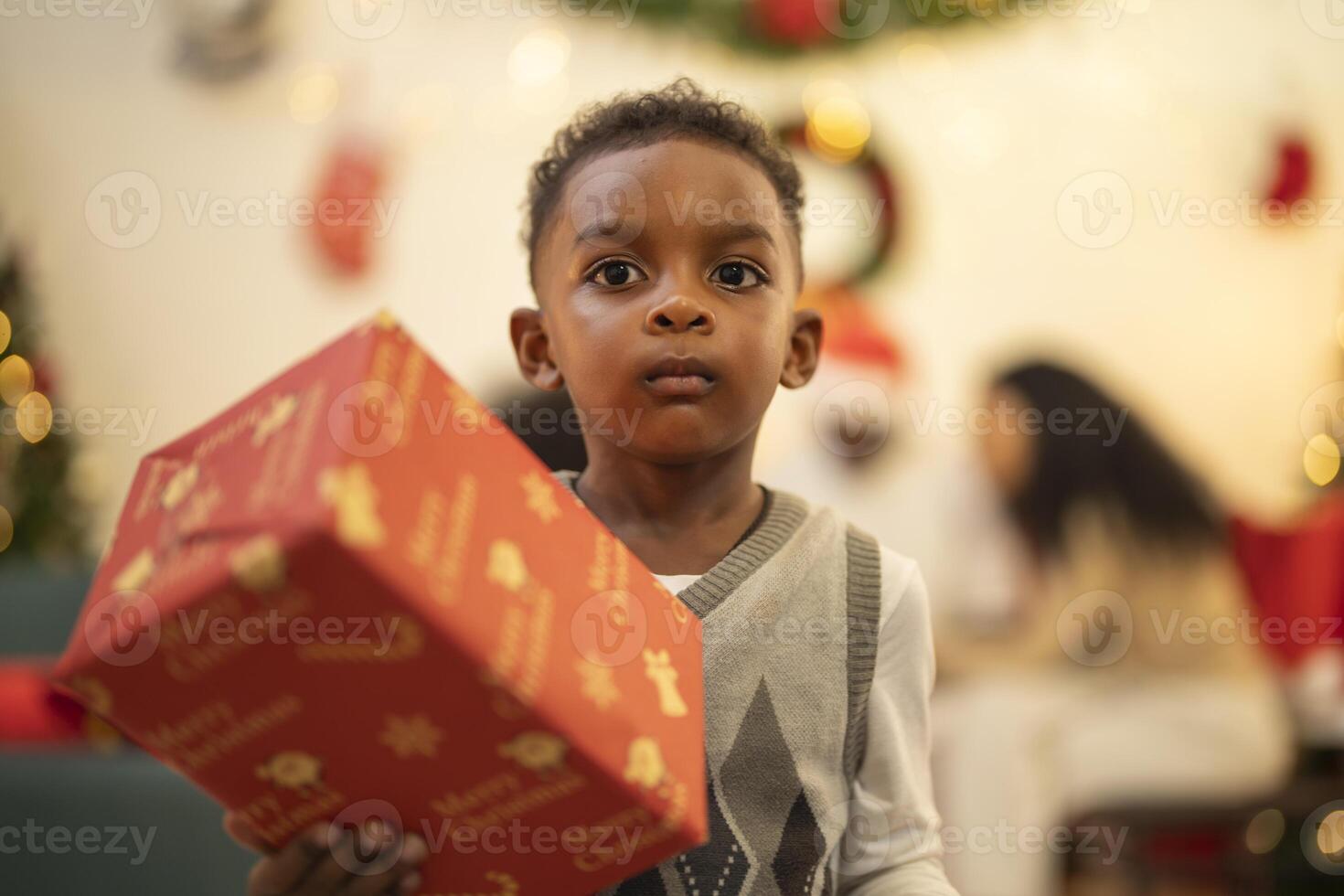 A young child excitedly holding a Christmas gift box, surrounded by festive decorations and twinkling lights in the background. The joyful expression on the child's face reflects the happiness. photo