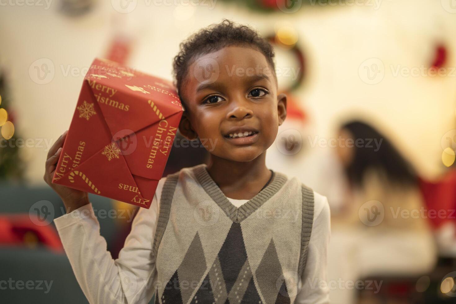 A young child excitedly holding a Christmas gift box, surrounded by festive decorations and twinkling lights in the background. The joyful expression on the child's face reflects the happiness. photo